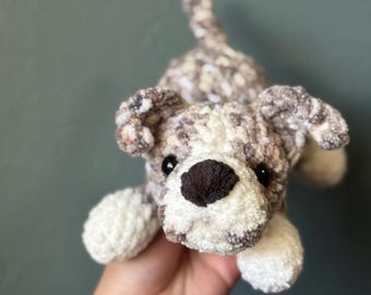 Puede incluir: Un pequeño perro de peluche tejido a mano con hilo jaspeado en tonos marrón, gris y blanco. El juguete tiene un hocico blanco sólido, una nariz de color marrón oscuro y dos ojos de plástico negro. Se muestra en una postura juguetona.