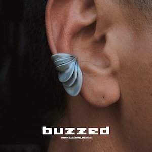 May include: A silver-colored ear cuff with a layered, petal-like design. The cuff is worn on the ear and is visible against the skin. The word "buzzed" is displayed in white, block letters, with "make some noise" below.