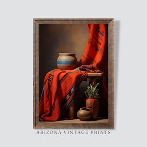 May include: A framed still life photograph featuring a terracotta pot with blue and white accents, a red and black patterned blanket draped over a wooden stool, and a small potted plant. The background is a muted brown.