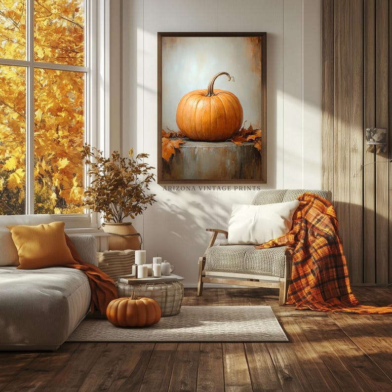 May include: A painting of a single orange pumpkin on a gray background with fall leaves. The painting is framed and hanging on a white wall in a living room setting. The room features a couch, a chair, and a coffee table with candles.
