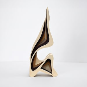 Puede incluir: Escultura abstracta de madera con un diseño en capas. La escultura presenta un exterior color crema con capas interiores marrones y negras. La forma es alta y fluida, con una base curva y una parte superior puntiaguda, creando una sensación de movimiento.