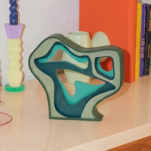 Peut inclure: Une sculpture abstraite en couches, dans des tons de bleu sarcelle, de bleu et de gris. L'œuvre d'art moderne a une forme organique unique avec des sections découpées. La sculpture est exposée sur une surface de couleur claire, avec des livres et d'autres objets décoratifs en arrière-plan.