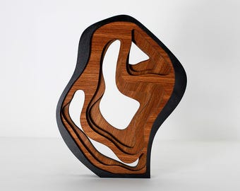 Op de afbeelding: Abstracte houten sculptuur met een gelaagd ontwerp. De sculptuur heeft een donkerbruine buitenkant met een zwarte rand en een lichtere bruine binnenkant met organische uitsparingen. Het kunstwerk wordt tegen een witte achtergrond gepresenteerd.