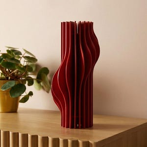 May include: A tall, red decorative vase with a unique, layered design. The vase has a curved, organic shape and is made of vertical, thin panels. It sits on a wooden surface, with a potted plant in the background.