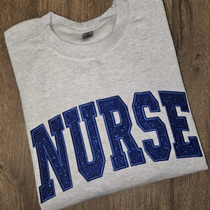May include: Gray sweatshirt with the word "NURSE" in large, blue, glittery letters.