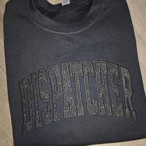 Puede incluir: Sudadera negra con la palabra "DISPATCHER" en letras de purpurina plateada.