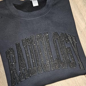 Op de afbeelding: Zwart sweatshirt met het woord "RADIOLOGY" in grote, glinsterende, zwarte letters.