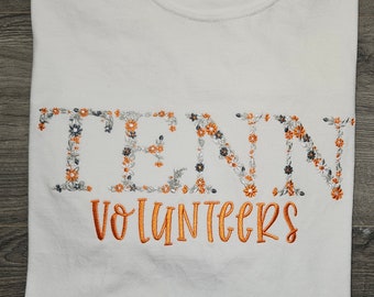 May include: White t-shirt with orange embroidery that says "TENNESSEE VOLUNTEERS". The letters are outlined with small gray and orange flowers.