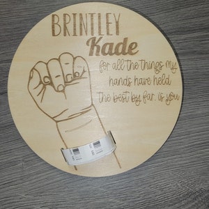 May include: A round wooden plaque with the names "Brintley" and "Kade" engraved in a decorative font. Below the names is a drawing of a hand making a fist. The text "for all the things my hands have held the best by far, is you" is also engraved.