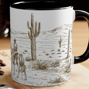Op de afbeelding: Witte keramische mok met een zwarte rand en handvat. De mok heeft een zwart-witte illustratie van een cowboy die op een paard door een woestijnlandschap met cactussen rijdt.