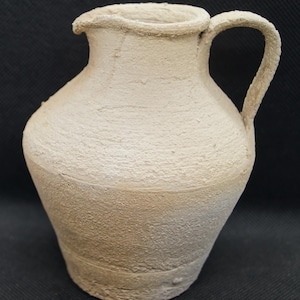 May include: A white ceramic jug with a rounded body and a single handle. The jug has a slightly textured surface and a slightly darker shade of white at the bottom.