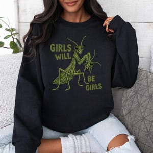 Pode incluir: Camisola preta com uma ilustração verde de dois louva-a-deus e o texto "GIRLS WILL BE GIRLS". O design é um gráfico caprichoso e único, perfeito para roupa casual.