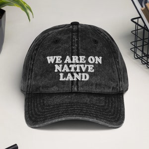 May include: Black vintage style baseball cap with white embroidered text that reads "We are on Native Land".
