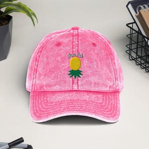 Può includere: Cappellino da baseball rosa con finitura invecchiata. Il cappellino presenta un ricamo di ananas con la scritta "Just Art" sopra. L'ananas è giallo con foglie verdi. Il cappellino è realizzato in materiale morbido e usurato.