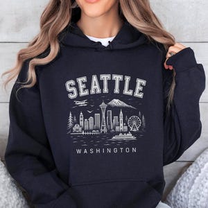 Puede incluir: Sudadera con capucha negra con un gráfico blanco del horizonte de Seattle, que incluye la Space Needle y una noria. La palabra "SEATTLE" está arqueada en la parte superior, con "WASHINGTON" debajo. La sudadera con capucha la lleva una persona.