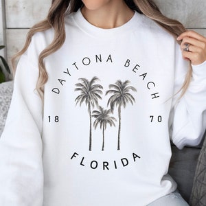 May include: White sweatshirt with black text that reads "Daytona Beach 1870 Florida" and a graphic of three palm trees.