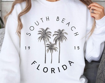 Peut inclure: Sweat-shirt blanc avec un motif noir de palmiers et le texte "South Beach Florida 19 15".
