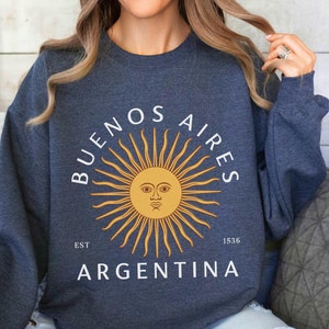 May include: A dark grey sweatshirt with a white graphic design featuring a stylized sun with a face, the text "BUENOS AIRES EST 1536 ARGENTINA", and a yellow sunburst.