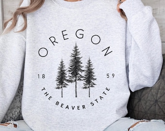 Può includere: Una felpa grigia con un design grafico nero di tre pini e il testo "OREGON 1859 THE BEAVER STATE".