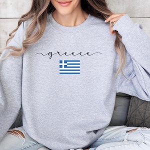 May include: A light gray sweatshirt with the word "greece" in a cursive font and a small Greek flag graphic. The flag is blue and white. The sweatshirt is a casual style.