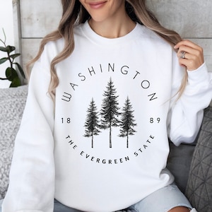 May include: White sweatshirt with a black graphic print of three evergreen trees in a circle. The text "WASHINGTON 1889 THE EVERGREEN STATE" is printed around the trees.