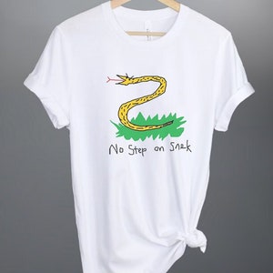 Puede incluir: Camiseta blanca con una ilustración de serpiente amarilla y el texto "No Step on Snek".