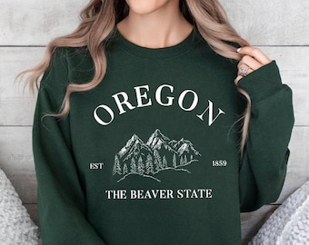 Può includere: Una felpa verde scuro con un design grafico bianco che dice "OREGON EST 1859 THE BEAVER STATE" con una catena montuosa e pini.