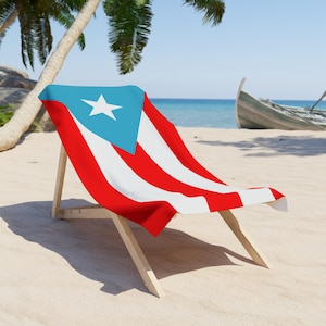 May include: A beach chair with a Puerto Rican flag draped over it. The flag features a white star on a blue triangle, with red and white stripes. The chair is made of light-colored wood and is on a sandy beach.