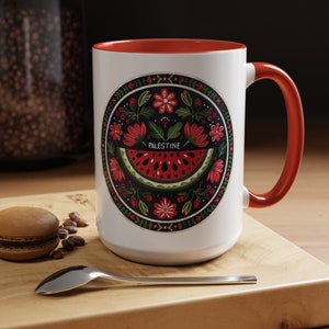 May include: White ceramic mug with a red rim and handle. The mug features a colorful illustration of a watermelon with the word "PALESTINE" in the center, surrounded by flowers and leaves.