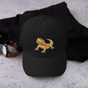 Op de afbeelding: Zwarte baseballcap met een geborduurd baardagamenmotief in oranje, geel en bruin. De cap heeft een gebogen klep en is gemaakt van een zacht materiaal. Op de achtergrond is een horloge met een bruine leren band te zien.