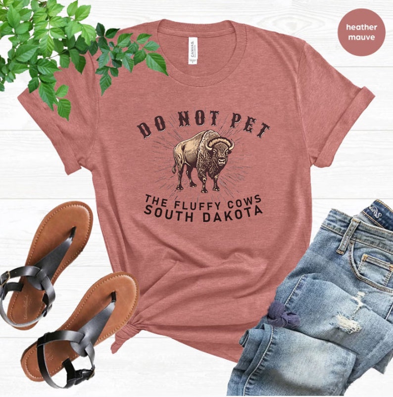 May include: A heather mauve t-shirt with the text "Do Not Pet The Fluffy Cows South Dakota" and a graphic of a brown bison.