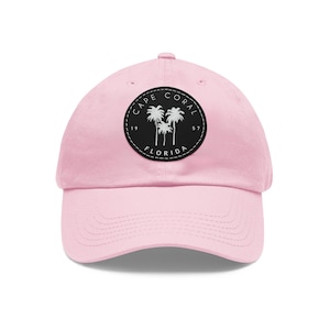 May include: A pale pink baseball cap with a curved peak. The cap has a circular black patch with the words "CAPE CORAL FLORIDA" and palm tree silhouettes in white. The hat is made of a soft fabric. The text includes the years 19 and 57.