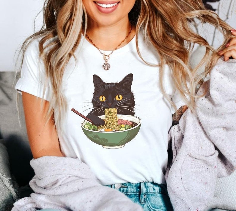 May include: White t-shirt featuring a black cat eating ramen from a green bowl. The cat has yellow eyes and is holding chopsticks. The ramen includes noodles, vegetables, and an egg. The shirt is a casual style.