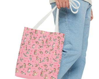 May include: Pink tote bag featuring a repeating pattern of gray and tan cats with red bows and yarn balls. The cats are in various poses. The bag has white straps and is held by a person wearing blue jeans.