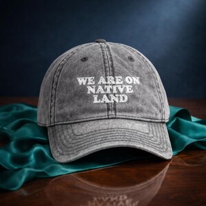 Può includere: Cappellino da baseball grigio con la scritta bianca "WE ARE ON NATIVE LAND". Il cappellino ha una visiera curva e un aspetto usurato. Lo sfondo presenta una superficie blu scuro e un tessuto verde acqua.