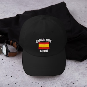 Puede incluir: Gorra de béisbol negra con texto bordado "Barcelona Spain" y un diseño de bandera roja y amarilla. La gorra está hecha de un material suave y tiene una visera curva. Un reloj con una correa de cuero marrón es visible en el fondo.