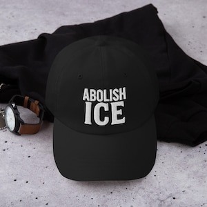 May include: Black baseball cap with the embroidered text "ABOLISH ICE" in white. The cap is made of a soft material and has a curved brim. A watch with a brown leather band is visible in the background.