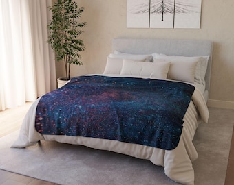 Puede incluir: Una manta de tiro azul y rosa con estampado de galaxia sobre una cama blanca con almohadas blancas.