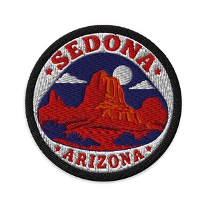 Puede incluir: Un parche bordado circular que muestra un paisaje desértico rojo con formaciones rocosas bajo un cielo azul oscuro con una luna blanca y nubes. El borde blanco contiene el texto rojo SEDONA en la parte superior y ARIZONA en la inferior.