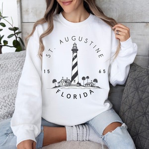 May include: White sweatshirt with a black and white illustration of a lighthouse, the text "ST. AUGUSTINE 1565 FLORIDA" and a palm tree design.