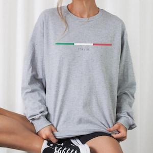 May include: A gray long-sleeve sweatshirt with a horizontal Italian flag design in green, white, and red. The word "ITALIA" is printed below the flag. The sweatshirt is a casual style.