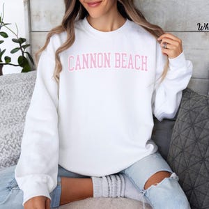 May include: A white sweatshirt with the text "CANNON BEACH" in pink block letters across the chest. The sweatshirt is a crew neck style and is being worn by a person. The person is wearing light blue jeans.
