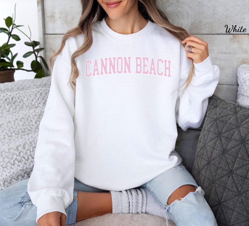 May include: A white sweatshirt with the text "CANNON BEACH" in pink block letters across the chest. The sweatshirt is a crew neck style and is being worn by a person. The person is wearing light blue jeans.