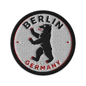 Peut inclure: Un écusson brodé circulaire représentant un ours noir debout sur ses pattes arrière au centre. Le fond est gris clair avec une fine bordure circulaire rouge. Le texte BERLIN est brodé en noir en haut et GERMANY en rouge en bas.