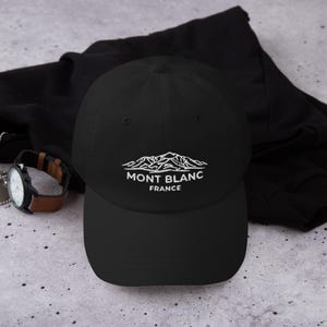 Peut inclure: Casquette de baseball noire avec le texte "MONT BLANC FRANCE" en blanc et un motif de chaîne de montagnes. La casquette est en matière souple et possède une visière incurvée. Une montre avec un bracelet en cuir marron est visible.