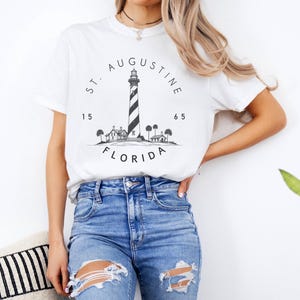 Può includere: T-shirt bianca con un'illustrazione in bianco e nero del faro di St. Augustine in Florida. Il design include il testo "ST. AUGUSTINE FLORIDA" e i numeri "15" e "65". La maglietta è abbinata a jeans blu.