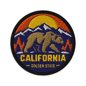 Puede incluir: Un parche bordado circular que muestra un oso pardo caminando frente a montañas blancas nevadas, pinos naranjas y un sol amarillo sobre un cielo azul oscuro. El texto CALIFORNIA y GOLDEN STATE está bordado en color amarillo.