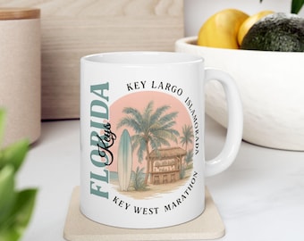 Puede incluir: Taza de cerámica blanca con una escena de playa con una tabla de surf, palmeras y una cabaña de playa. La taza tiene las palabras "Florida Keys" y los nombres de varias islas.