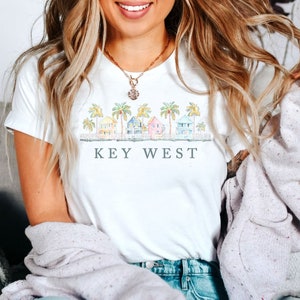 May include: White t-shirt featuring a watercolor illustration of colorful houses and palm trees. The text "KEY WEST" is printed below the image. The shirt is made of a soft material.
