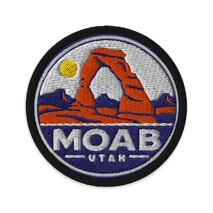 Può includere: Una toppa ricamata circolare che raffigura il paesaggio dell'arancione Delicate Arch a Moab, nello Utah, su un cielo grigio chiaro con un sole giallo. La parte inferiore è blu scuro con il testo bianco MOAB e UTAH sotto l'arco.